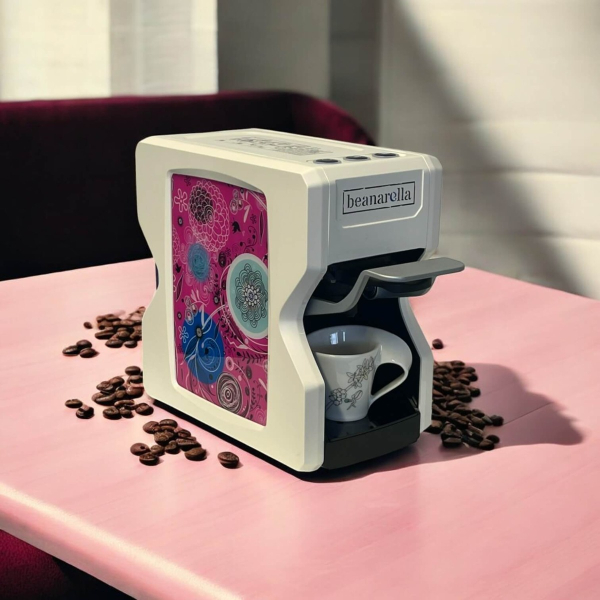 Machine à café Nori Édition Spéciale - Floral Dream, blanche avec un design floral rose. Compatible avec des capsules compostables, des dosettes et des capsules rechargeables. Des grains de café sont disposés sur la table avec une tasse sous le bec verseu