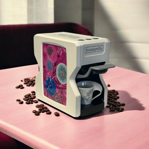 Macchina da caffè Nori Edizione Speciale - Floral Dream, bianca con design floreale rosa. Compatibile con capsule compostabili, cialde e capsule ricaricabili. Chicchi di caffè sono sparsi sul tavolo, con una tazzina sotto il beccuccio.