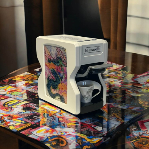 Macchina da caffè Nori Edizione Speciale – Design Drago. Macchina da caffè bianca con un motivo artistico di drago cinese su una superficie colorata. Una tazza è posta all'interno della macchina e chicchi di caffè sono sparsi sul tavolo.