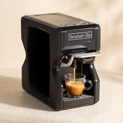 Macchina da caffè a capsule Nori Nero di beanarella che eroga un espresso in una tazzina di vetro – macchina nera con tre tasti, vista di tre quarti
