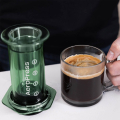 AeroPress Coffee Maker - Clear Green à côté d’une tasse de café fraîchement préparé.


