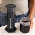 AeroPress Coffee & Espressomaker - XL placée à côté d'une carafe contenant du café fraîchement préparé, avec une personne partiellement visible en arrière-plan.


