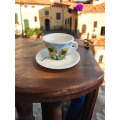 Tasse à café Aquarelli avec un motif de tournesol sur une table en bois devant un village italien.


