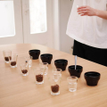 Hario Cupping Bowl Kasuya, présenté sur une table de dégustation avec des verres et des grains de café.


