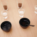 Cuillère de cupping Hario Kasuya sur une table en bois avec des tasses noires et des verres pour une dégustation.


