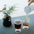 Thermopot Hario 600 ml versant du café sans gouttes dans un verre, décoré avec une plante à côté.

