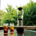 Slow Drip Brewer HARIO SHIZUKU de 600 ml, placé sur une table de jardin avec deux verres remplis.


