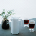 Hario Thermopot 600 ml en blanc sur une table avec deux verres de café fraîchement préparé et une plante en arrière-plan.

