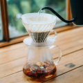 Kit Hario V60 Glass Brewing préparant un café en méthode Pour-Over sur une table en bois, vue sur une fenêtre.


