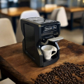 Nori black coffee machine, angled on a wooden table, with coffee beans and a cup under the spout.


