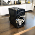 Nori black coffee machine on a wooden countertop in a modern kitchen, with a cup placed under the spout ready for coffee.


