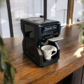 Beanarella Nori black coffee machine, placed on a wooden counter, with a cup under the spout. Modern environment with natural accents.


