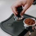 Rhinowares Tamper-Matte mit abgewinkelter Kante – Tamper wird neben einem mit Kaffeemehl gefüllten Siebträger auf die Matte gestellt

