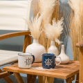 Tasses en céramique au design nautique en Bleu et Blanc, décorées avec des symboles de barre de gouvernail et ancre, parfaites pour une pause café élégante. Idéalement placées sur une table en bois, à côté de vases blancs modernes avec des graminées de pa