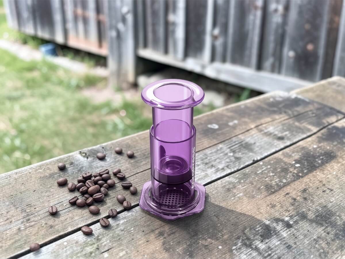 AeroPress Coffee Maker Clear Purple on rustic wooden bench with coffee beans outdoors