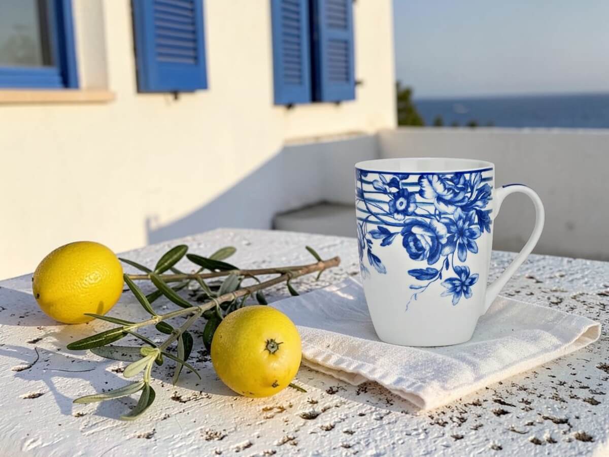 Porcelain mug Blaue Blütenranke with blue floral pattern on linen cloth on white stone terrace with lemons olive branch and blue shutters with sea view in sunlight
