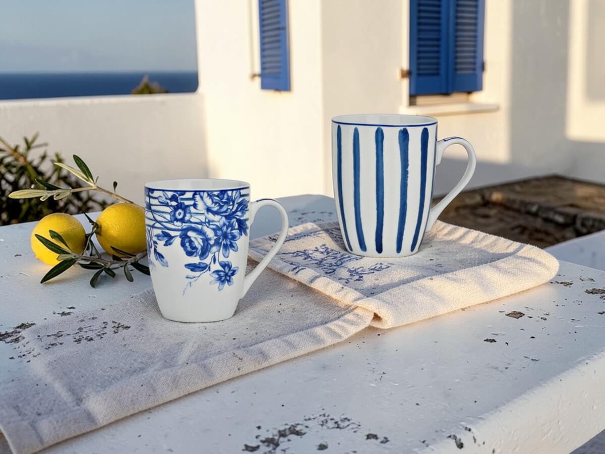 Two porcelain mugs Blaue Stunde with floral pattern and stripes on linen cloth on white stone terrace with lemons olive branch and blue shutters in Greek style with sea view