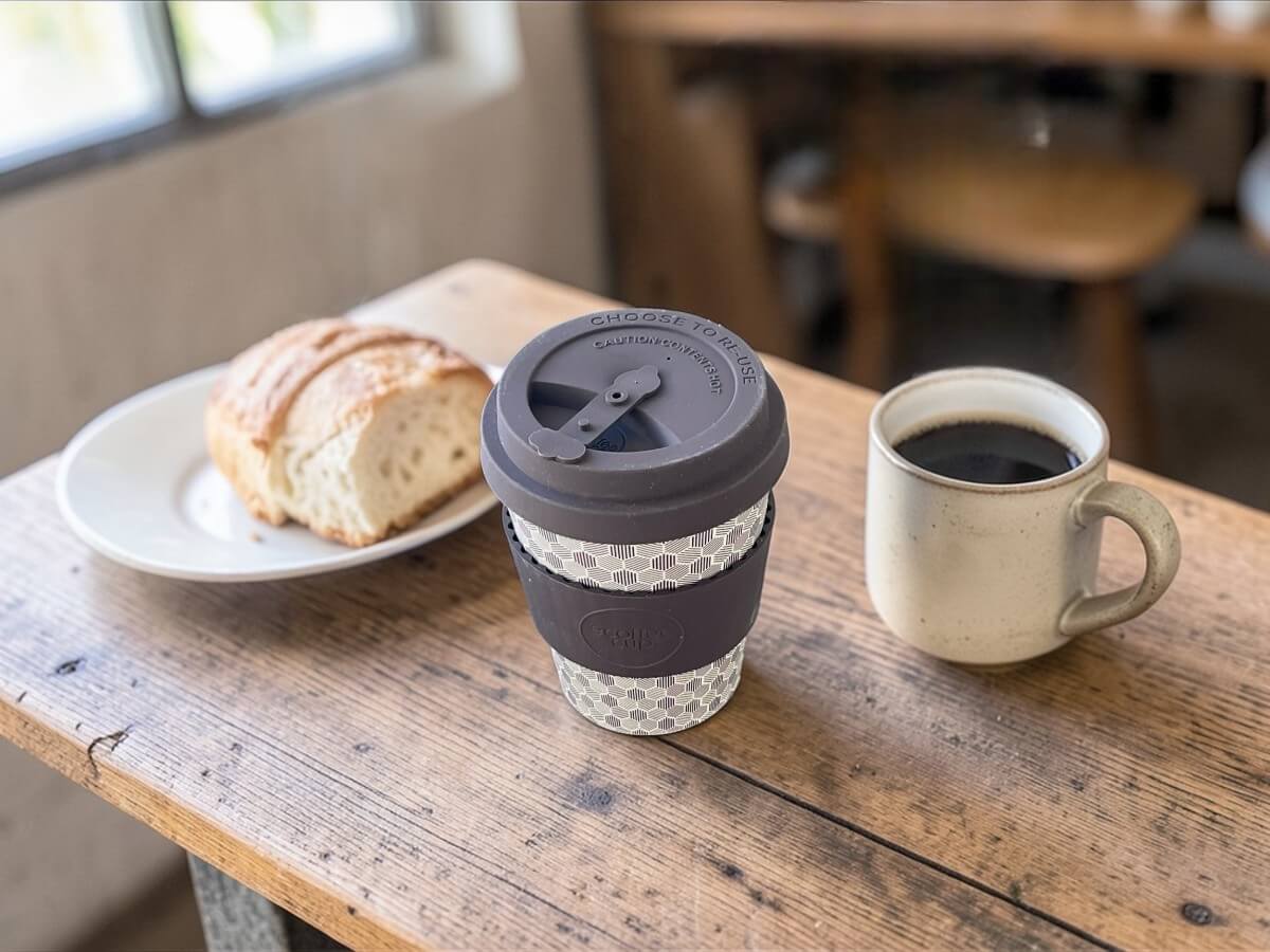 Ecoffee Cup bicchiere coffee to go 350ml Fermis Paradox in grigio scuro con motivo geometrico cubico su tavolo in legno rustico con pane fresco su piatto in ceramica e tazza da caffè con caffè nero in caffetteria accogliente