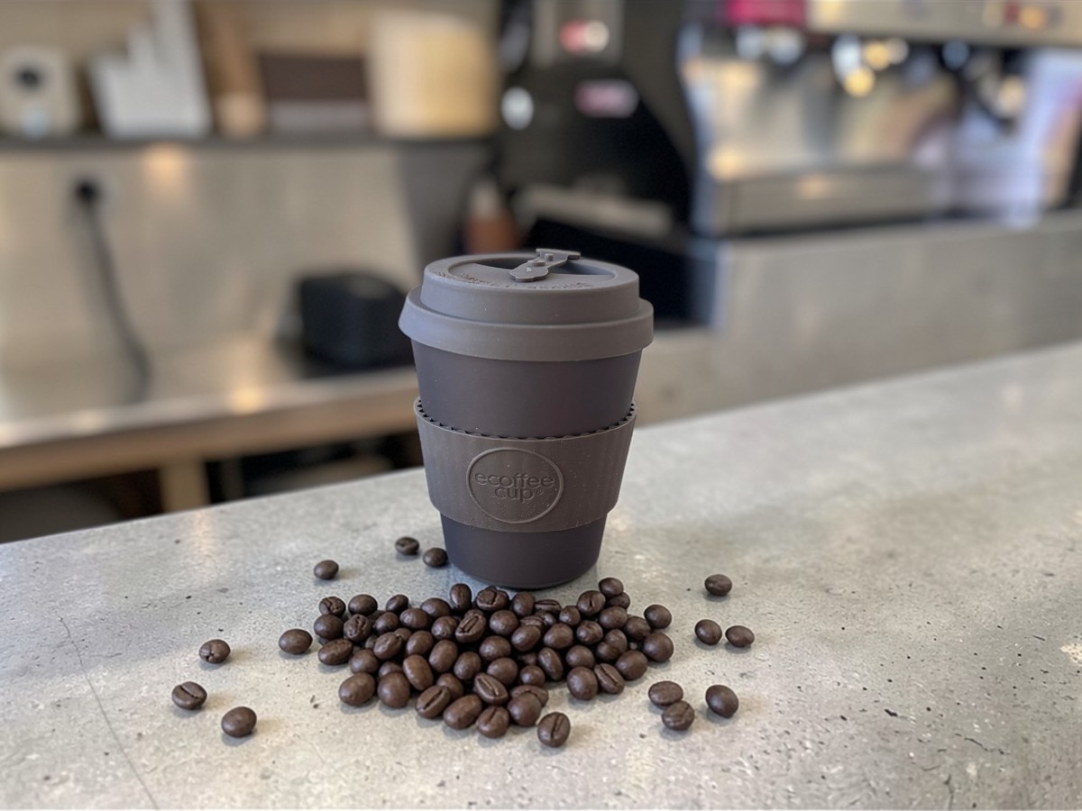 Ecoffee Cup Espressobecher Mokka Braun 180ml Spooky Action At A Distance auf grauer Betontheke mit verstreuten Kaffeebohnen und professioneller Espressomaschine im Hintergrund in Kaffeebar-Atmosphäre