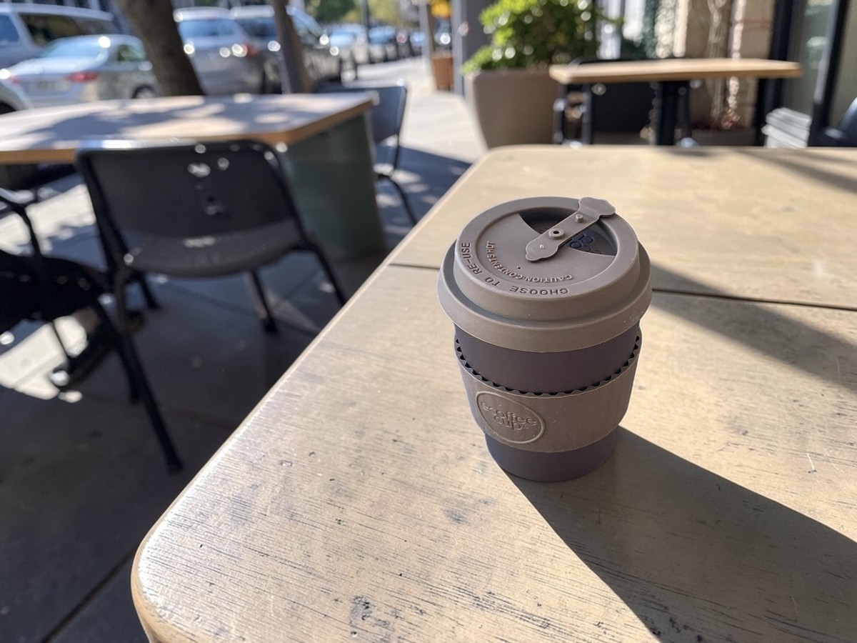 Ecoffee Cup Espressobecher Mokka Braun 180ml Spooky Action At A Distance auf Holztisch einer Café-Terrasse im Sonnenlicht mit Metallstühlen und Stadtszene im Hintergrund