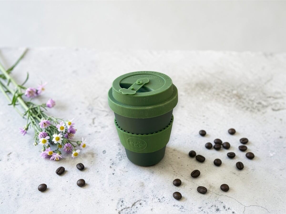 Ecoffee Cup Espressobecher Olivenmoos Grün 180ml Oberon auf heller Betonoberfläche neben lila Blüten und verstreuten Kaffeebohnen im natürlichen Tageslicht