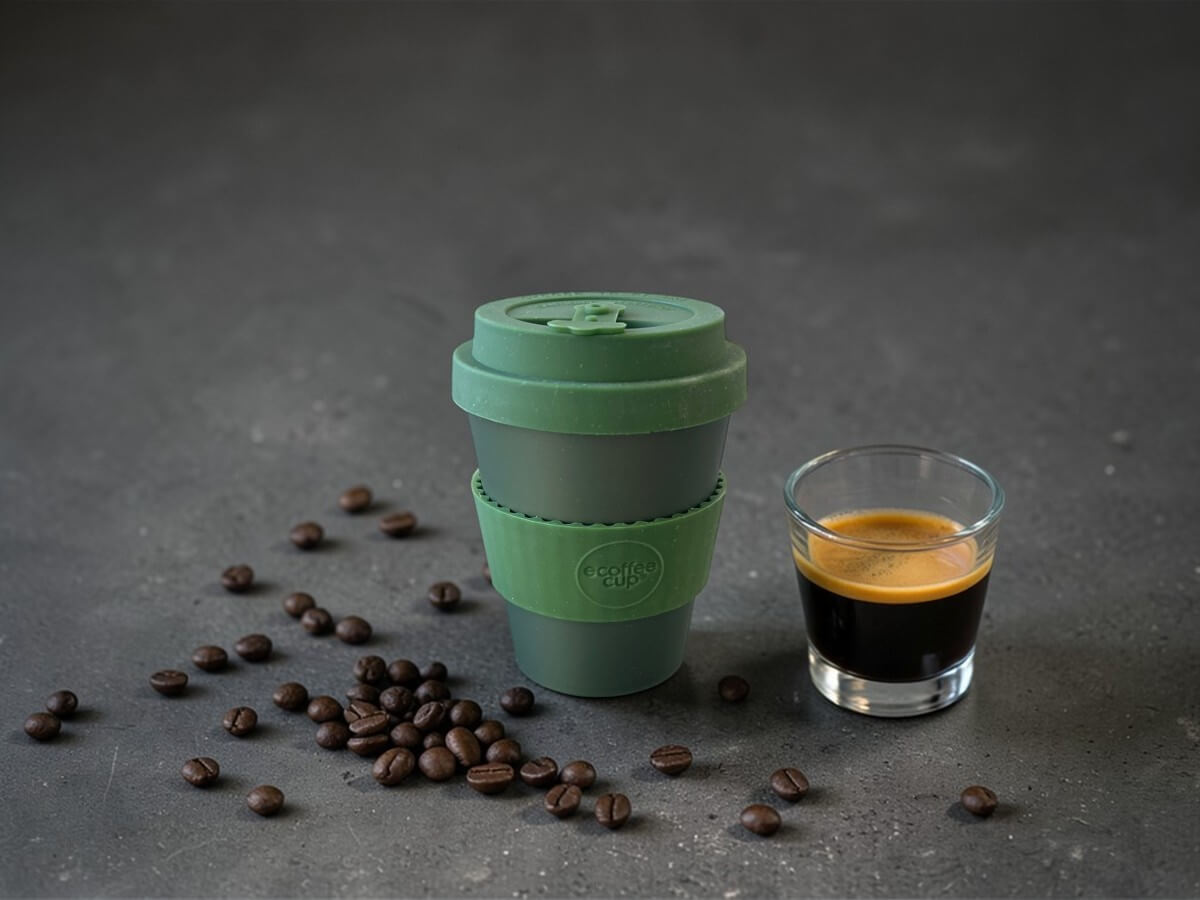 Ecoffee Cup Espressobecher Olivenmoos Grün 180ml Oberon auf dunkler Betonoberfläche neben Espresso im Glas mit Crema und verstreuten Kaffeebohnen in stimmungsvoller Beleuchtung