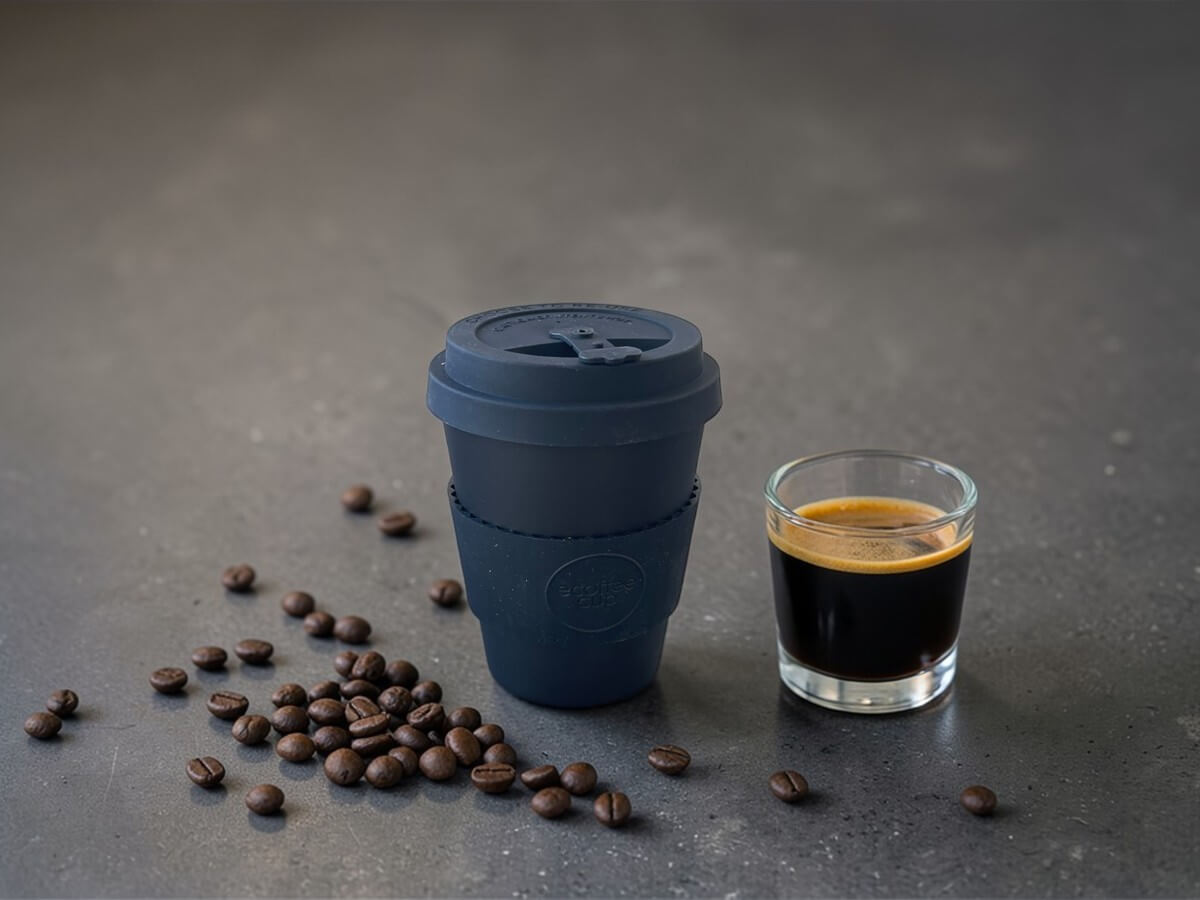 Ecoffee Cup Espressobecher Schwarz 180ml Kerr & Napier auf dunkler Betonoberfläche neben Espresso im Glas mit Crema und verstreuten Kaffeebohnen in stimmungsvoller Beleuchtung