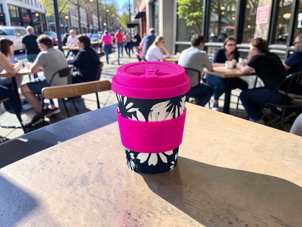 Ecoffee Cup Inflorescence 350ml in rosa con motivo floreale bianco e nero su tavolo in legno di terrazza caffetteria animata con clienti e vita urbana nella luce estiva