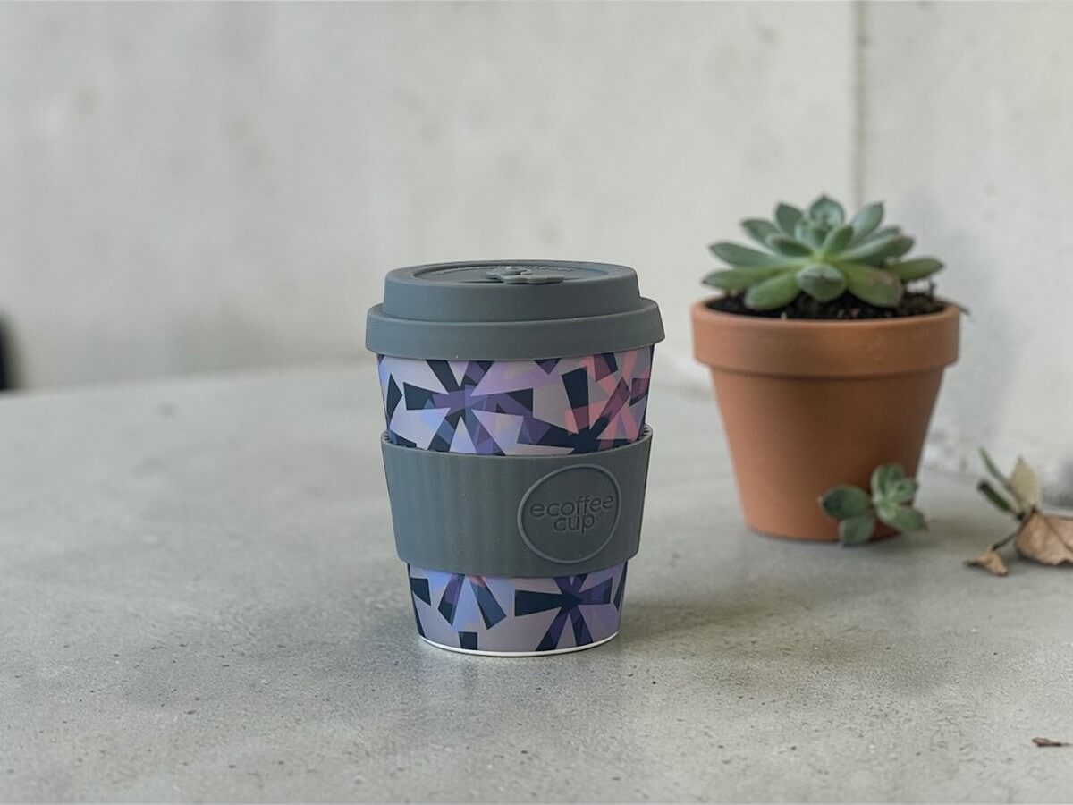 Ecoffee Cup coffee cup 240ml Dunning Kruger in grey with geometric crystal pattern in purple pink and blue on light concrete surface next to succulent in terracotta pot against concrete wall