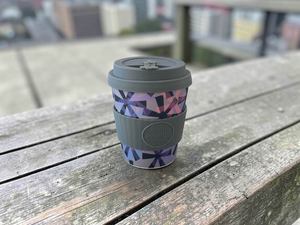 Ecoffee Cup coffee cup 240ml Dunning Kruger in grey with geometric crystal pattern in purple pink and blue on weathered wooden bench with balcony railing and city view in daylight
