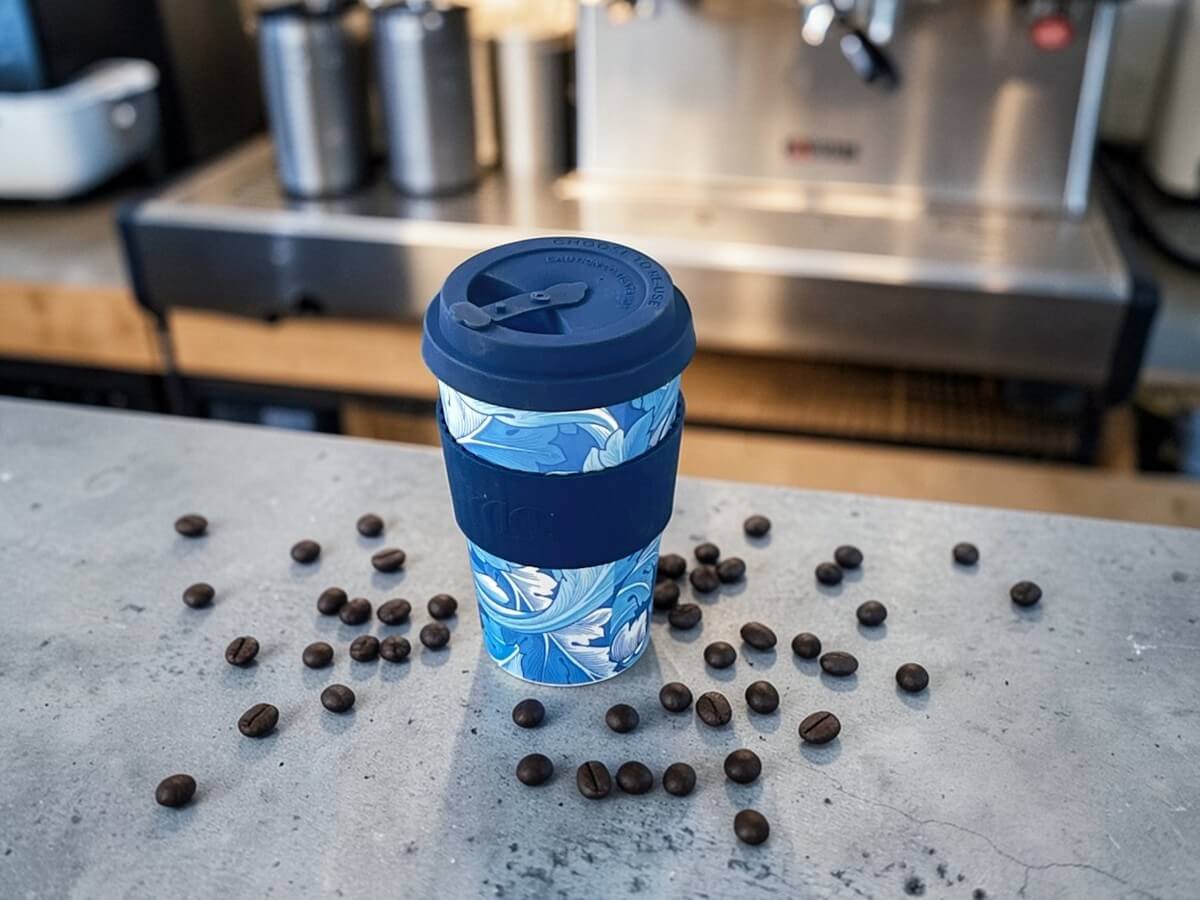 Ecoffee Cup bicchiere riutilizzabile 400ml WMG Acanthus in blu con motivo floreale a foglie di acanto su bancone in cemento con chicchi di caffè sparsi e macchina espresso professionale in acciaio inossidabile sullo sfondo