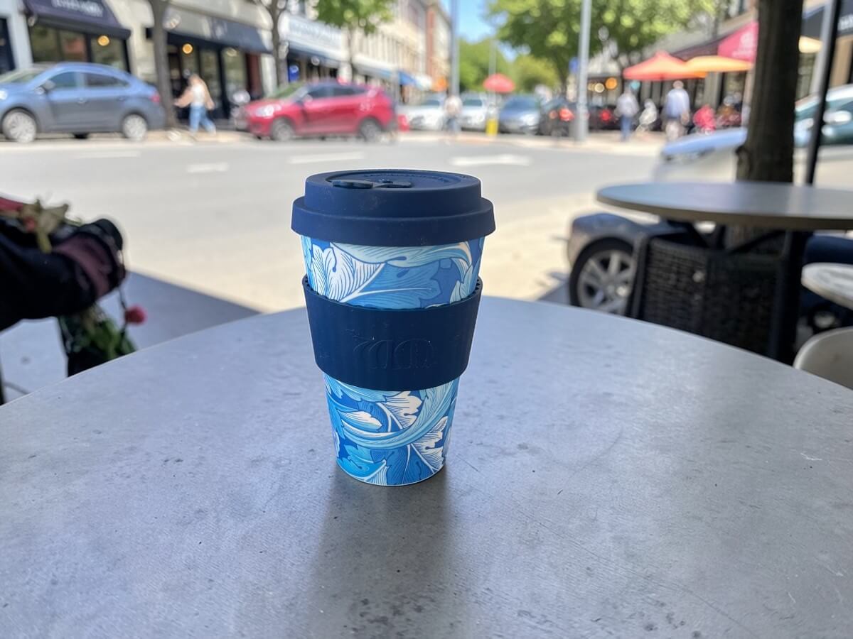 Ecoffee Cup bicchiere riutilizzabile 400ml WMG Acanthus in blu con motivo floreale a foglie di acanto su tavolo rotondo in metallo di terrazza caffetteria con strada urbana animata e sole d'estate