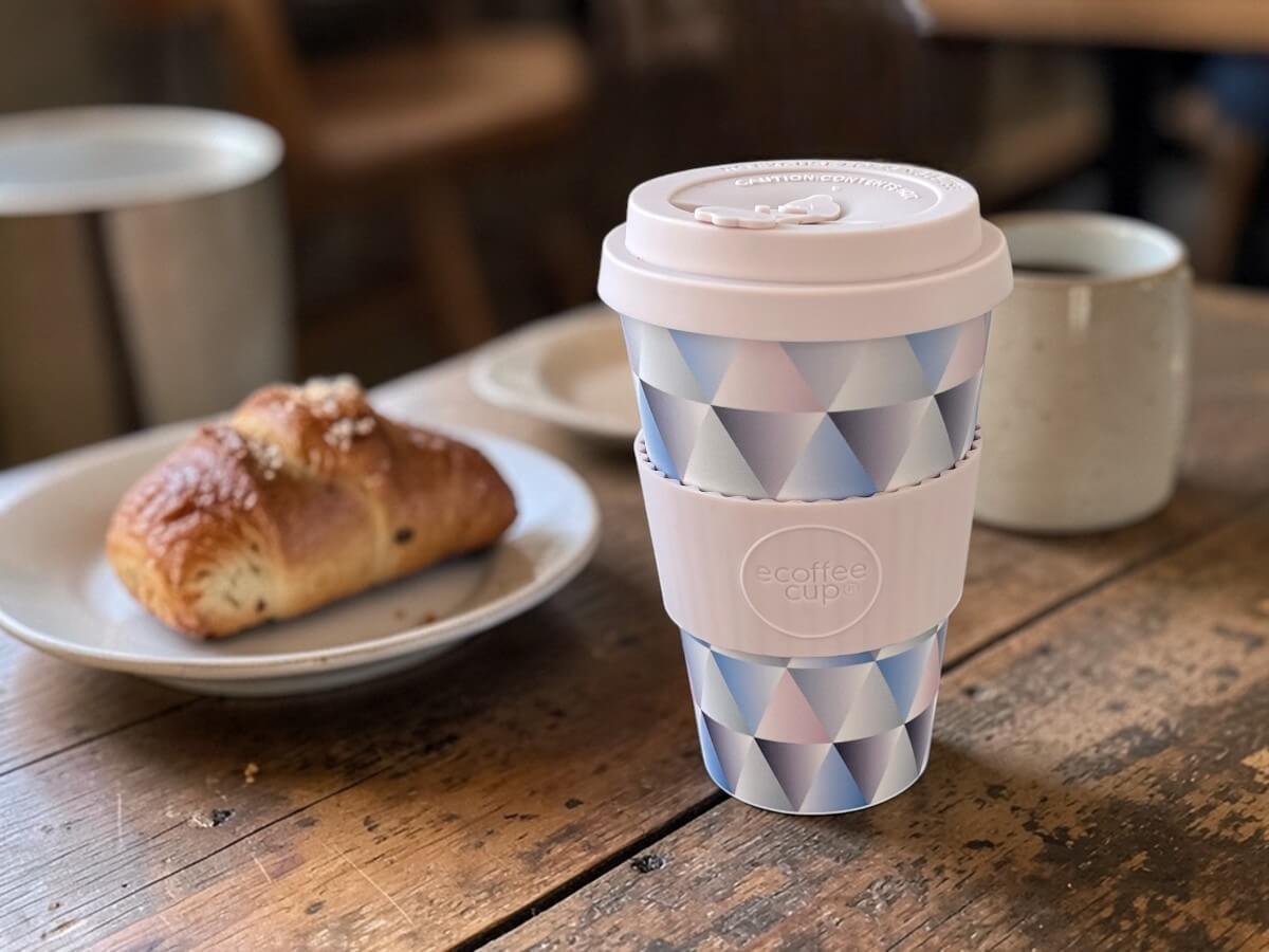Ecoffee Cup Mehrwegbecher Pastell-Mosaik 400ml Shandor the Magnificent mit geometrischem Dreiecksmuster in Pastelltönen auf rustikalem Holztisch mit Croissant auf Keramikteller und Kaffeetasse im gemütlichen Café-Frühstückssetting