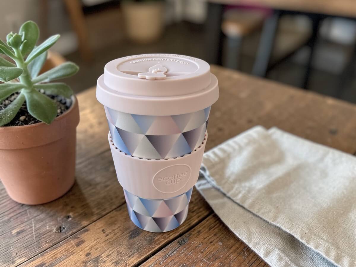 Ecoffee Cup Mehrwegbecher Pastell-Mosaik 400ml Shandor the Magnificent mit geometrischem Dreiecksmuster in Pastelltönen auf rustikalem Holztisch mit Sukkulente im Terrakotta-Topf und Leinenbeutel in gemütlichem Café