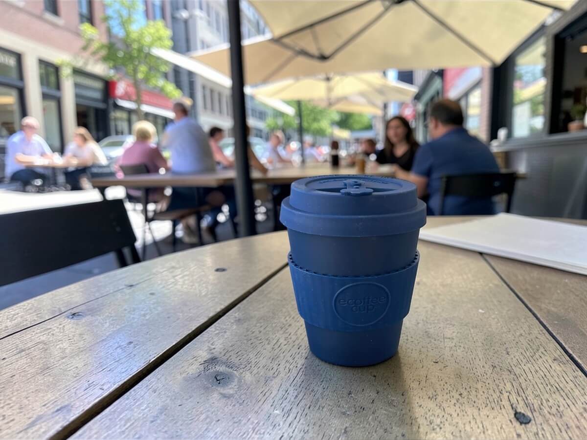 Ecoffee Cup bicchiere da caffè sostenibile 240ml Dark Energy in blu scuro su tavolo in legno di terrazza caffetteria animata con ombrelloni e clienti nella vita urbana estiva