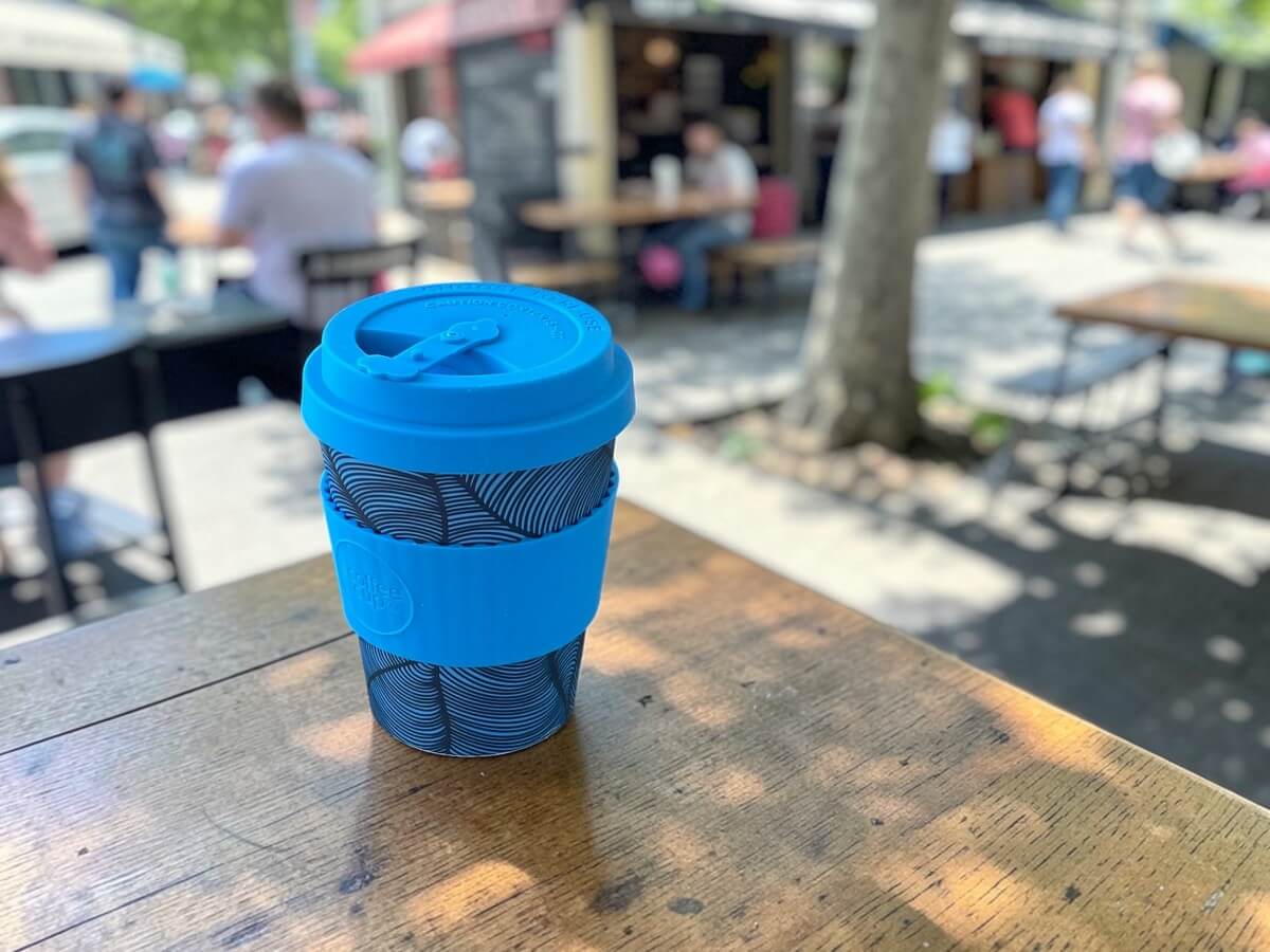 Ecoffee Cup bicchiere da caffè sostenibile 350ml Dotonbori in blu con motivo ondulato su tavolo in legno di terrazza caffetteria all'ombra di un albero con scena urbana animata in una giornata estiva