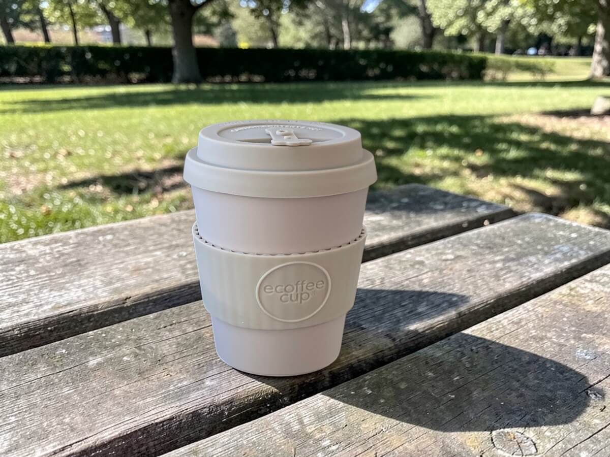 Ecoffee Cup bicchiere da caffè sostenibile 350ml Molto Grigio su panchina da parco in legno invecchiato con prato verde e alberi alla luce del sole sullo sfondo