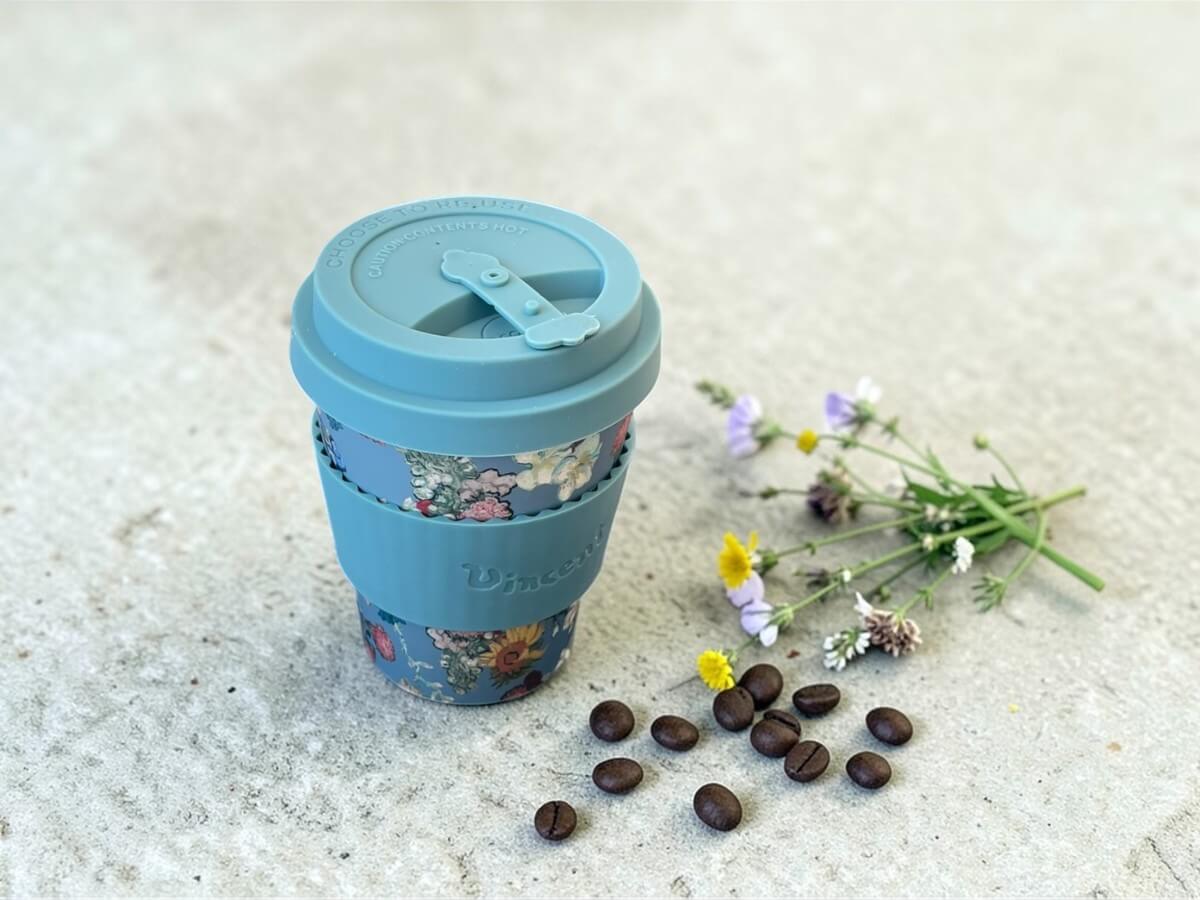 Ecoffee Cup bicchiere da caffè sostenibile 350ml VGM 50th Anniversary in blu colomba con motivo bouquet floreale e scritta Vincent su superficie in cemento con fiori selvatici e chicchi di caffè alla luce naturale