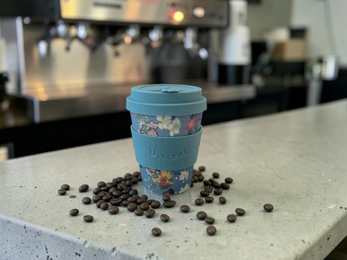 Ecoffee Cup bicchiere da caffè sostenibile 350ml VGM 50th Anniversary in blu colomba con motivo bouquet floreale e scritta Vincent su bancone in cemento con chicchi di caffè sparsi e macchina espresso professionale sullo sfondo