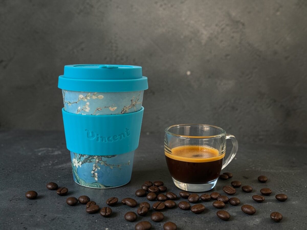 Ecoffee Cup nachhaltiger Kaffeebecher 350ml VGM Almond Blossom in Türkis mit Mandelblüten-Motiv und Vincent-Schriftzug auf dunkler Betonoberfläche neben Espresso im Glas mit Crema und verstreuten Kaffeebohnen