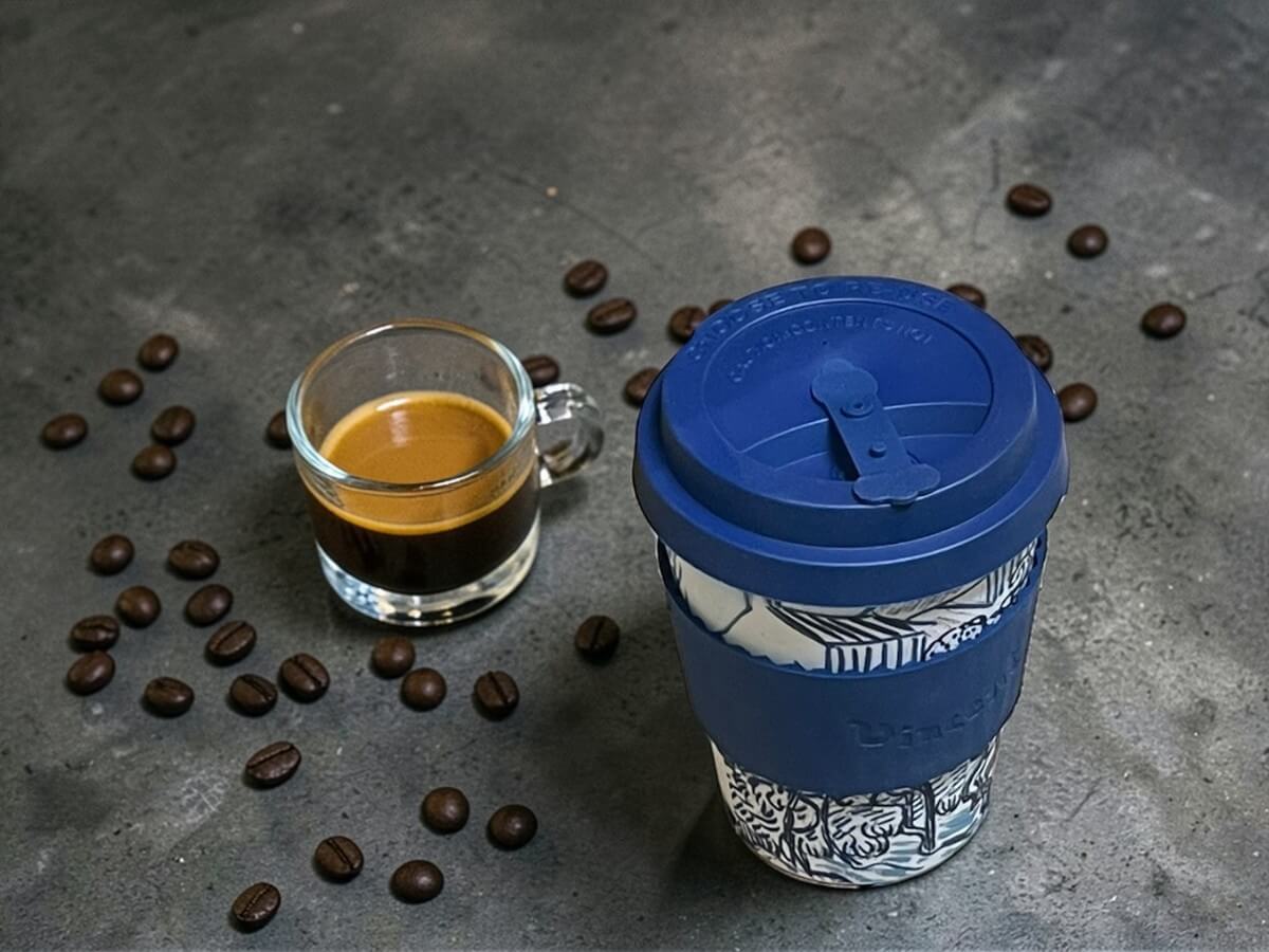 Ecoffee Cup bicchiere da caffè sostenibile 350ml VGM Old Vineyard with Peasant Woman in blu scuro con motivo vigneto e scritta Vincent su superficie in cemento scuro accanto a espresso in vetro con crema e chicchi di caffè sparsi