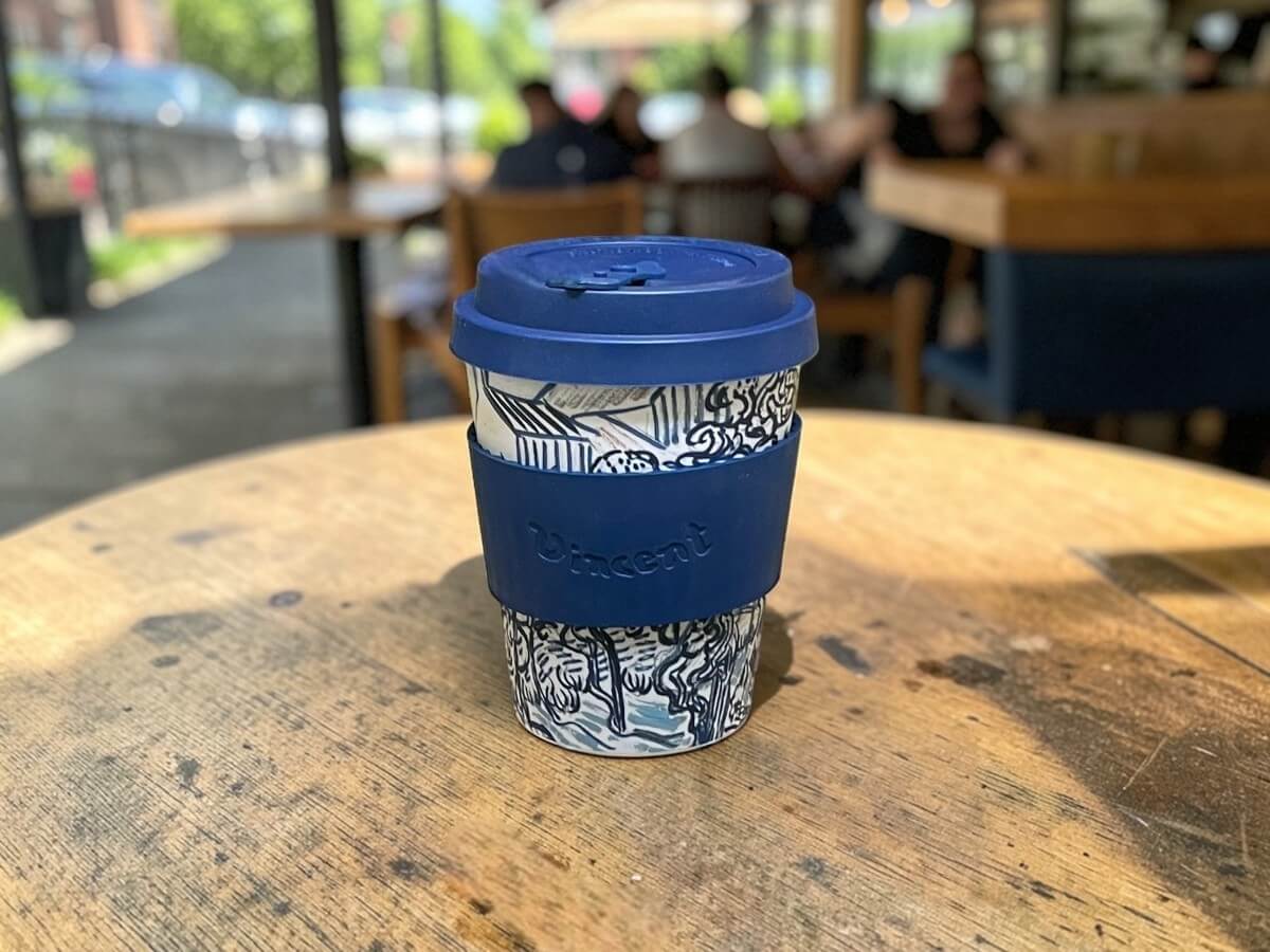 Ecoffee Cup bicchiere da caffè sostenibile 350ml VGM Old Vineyard with Peasant Woman in blu scuro con motivo vigneto e scritta Vincent su tavolo rotondo in legno di terrazza caffetteria con clienti nella luce estiva