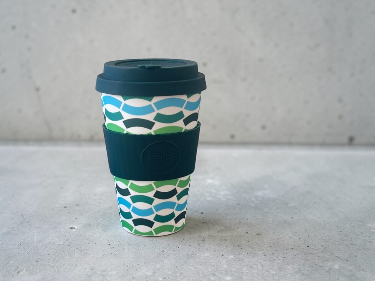 Ecoffee Cup bicchiere da caffè sostenibile 400ml Bloki Balentina in verde scuro con motivo geometrico ondulato in verde blu e bianco su superficie in cemento davanti a parete in cemento in ambientazione minimalista