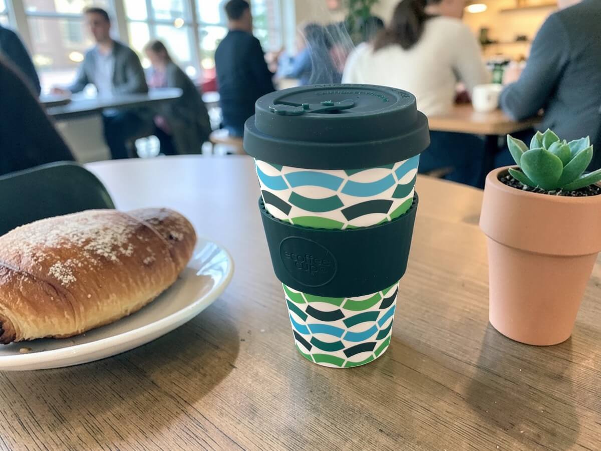 Ecoffee Cup bicchiere da caffè sostenibile 400ml Bloki Balentina in verde scuro con motivo geometrico ondulato in verde blu e bianco su tavolo in legno di caffetteria accanto a croissant al cioccolato e succulenta in vaso con clienti sullo sfondo