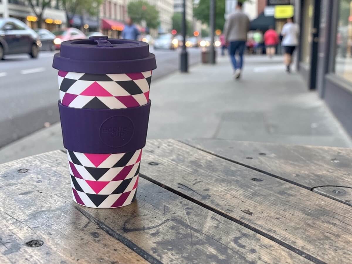 Ecoffee Cup nachhaltiger Kaffeebecher 400ml Fancy Wang in Lila mit geometrischem Dreiecksmuster in Pink Schwarz und Weiss auf Holztisch einer Café-Terrasse mit Passanten und Stadtszene im Abendlicht
