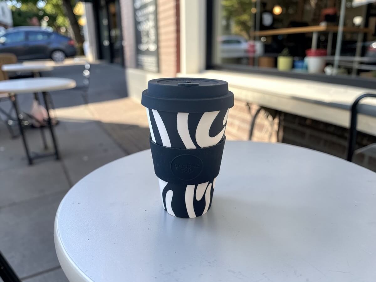 Ecoffee Cup bicchiere da caffè sostenibile 400ml Manassa's Run in nero con motivo ondulato astratto bianco su tavolo rotondo in metallo di terrazza caffetteria con scena urbana alla luce del sole