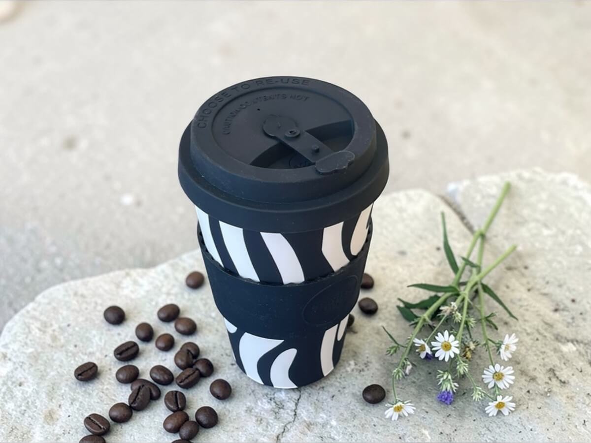 Ecoffee Cup bicchiere da caffè sostenibile 400ml Manassa's Run in nero con motivo ondulato astratto bianco su superficie in pietra con chicchi di caffè sparsi e fiori selvatici alla luce naturale