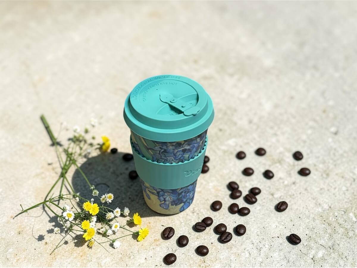 Ecoffee Cup sustainable coffee cup 400ml VGM Irises in green with irises motif and Vincent lettering on light concrete surface next to chamomile flowers and scattered coffee beans in sunlight