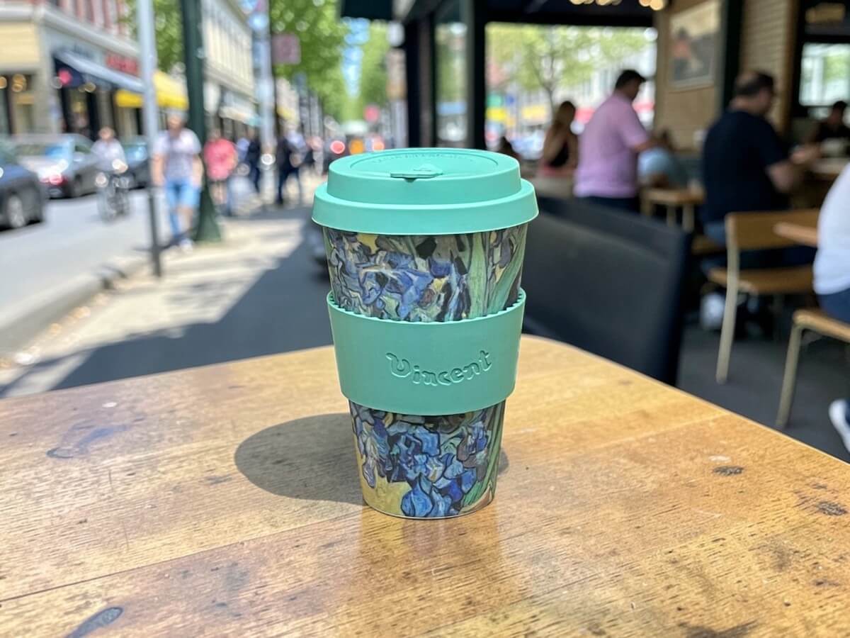 Ecoffee Cup sustainable coffee cup 400ml VGM Irises in green with irises motif and Vincent lettering on wooden cafe terrace table with busy city scene and guests in summer sunlight