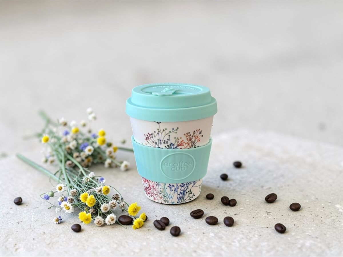 Ecoffee Cup bicchiere to go sostenibile 240ml Little Canning in verde menta con motivo a rametti colorati su superficie in cemento chiaro accanto a mazzolino di fiori selvatici e chicchi di caffè sparsi alla luce naturale